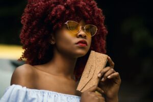 pexels-godisable-jacob-226636-1191520 (1) A stylish woman with voluminous red curls, wearing yellow-tinted cat-eye sunglasses and an off-shoulder top, confidently holding a cork-textured clutch