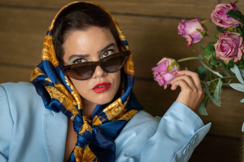 pexels-israyosoy-27155382 A stylish woman wearing a blue and gold headscarf, dark cat-eye sunglasses, and a light blue blazer, holding a pink rose near her face against a wooden background.