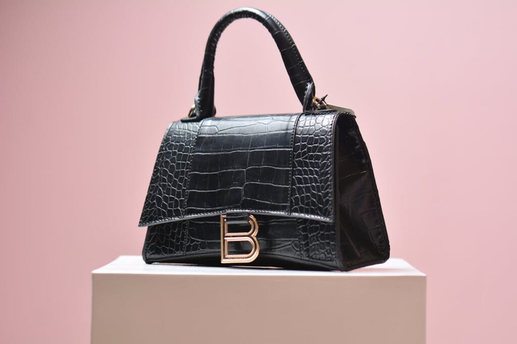 pexels-tarek-shahin-153182669-12428373 A black crocodile-embossed handbag with a structured design, gold-tone "B" logo clasp, and a top handle, displayed on a white pedestal against a soft pink background.