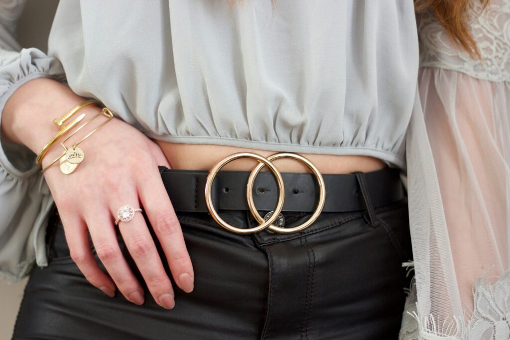 thea-hdc-cvJySBfdyOw-unsplash (1) Close-up of a woman's waist showcasing a black belt with a gold double-ring buckle, paired with a light gray blouse and black leather pants, accessorized with gold bracelets and a statement ring.