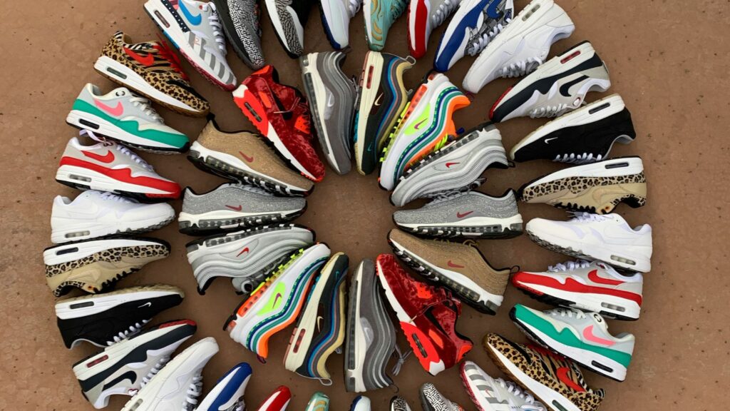 A visually striking circular arrangement of various Nike Air Max sneakers in different colors, patterns, and textures, including leopard print, metallic, bright neon, and classic white designs, displayed on a brown surface.