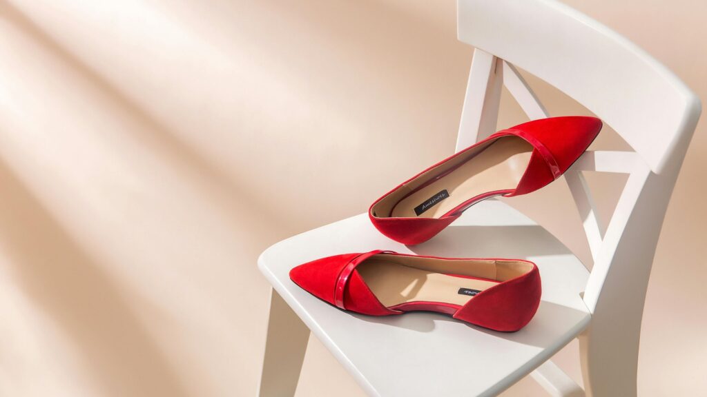A pair of elegant red suede pointed-toe flats with glossy details, carefully placed on a white wooden chair against a softly lit beige background, casting gentle shadows.