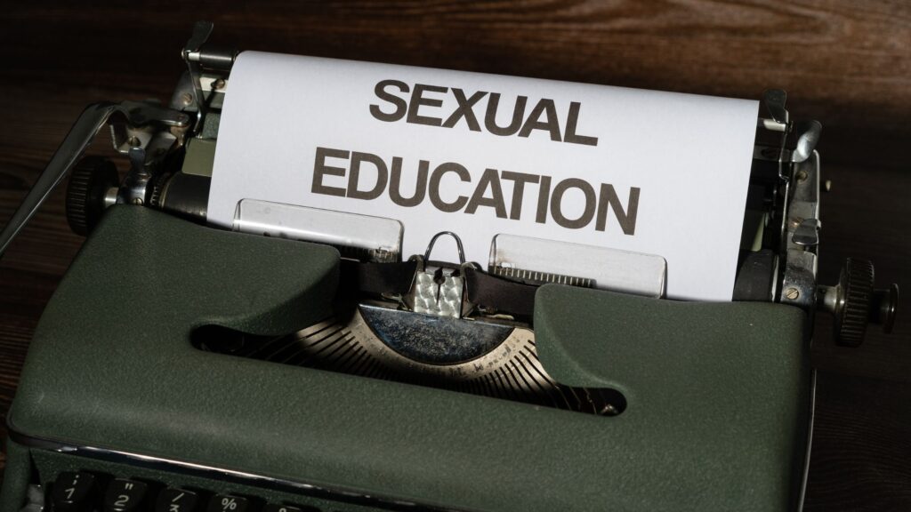 A vintage green typewriter with a sheet of white paper inserted, displaying the bold, black words 'SEXUAL EDUCATION.' The background features a wooden surface.