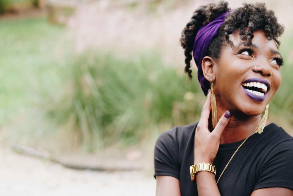 A joyful young woman with dark, curly hair styled in a purple headband smiles brightly. She wears bold purple lipstick, gold jewelry, and a black top. Her hand gently touches her neck as she looks away, with a blurred outdoor background of greenery.