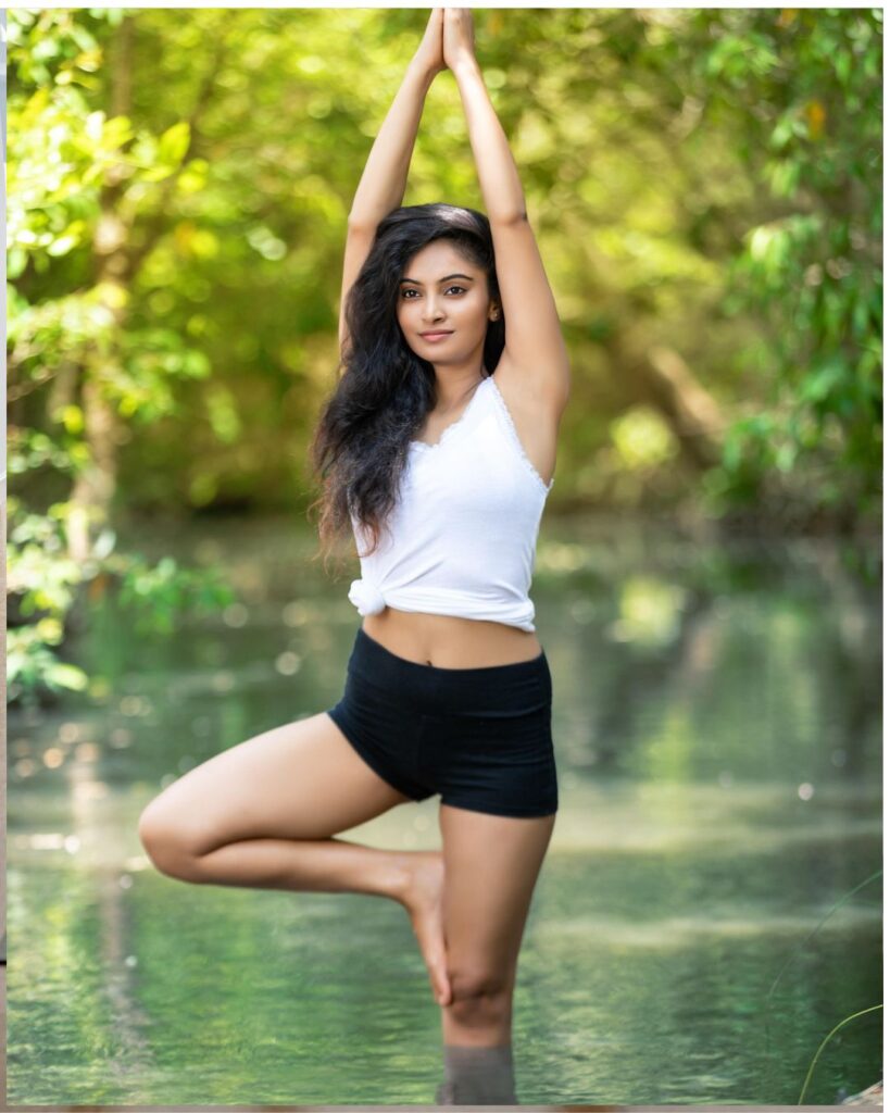 A young woman with long, wavy hair practices yoga outdoors, standing in a tree pose with one foot on her inner thigh and arms raised above her head. She wears a white sleeveless top and black shorts, balancing gracefully in a shallow body of water surrounded by lush green trees.