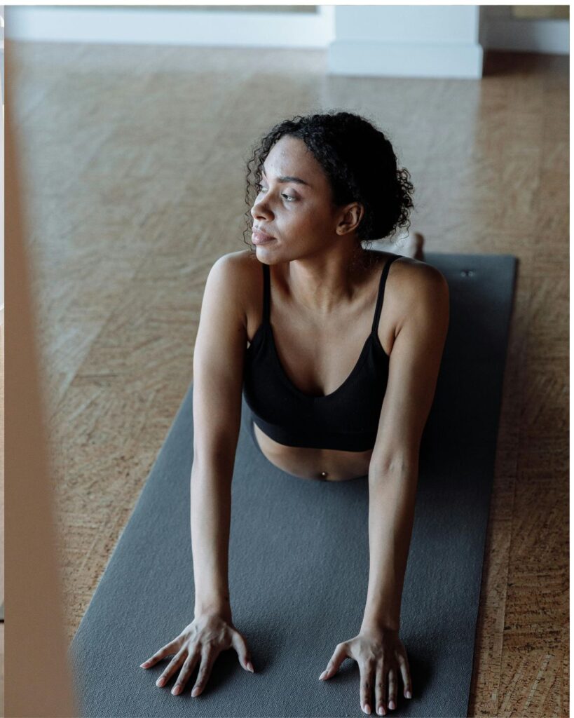 A young woman with curly hair practices yoga indoors, performing the cobra pose on a gray yoga mat. She wears a black sports bra and gazes to the side with a calm and focused expression. The room has a wooden floor and soft natural lighting.