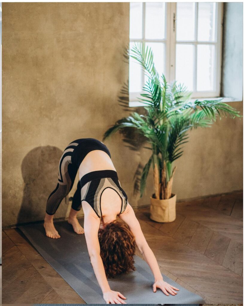 A woman with curly hair practices yoga indoors, performing the downward-facing dog pose on a gray yoga mat. She wears a black and gray athletic outfit. The room has wooden flooring, a textured beige wall, a large window, and a potted plant in the background.