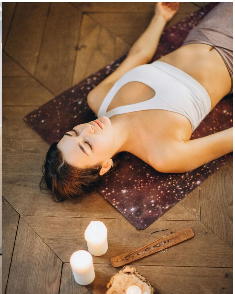 A woman in a white sports top and gray leggings lies on her back on a yoga mat with a starry design, practicing the Savasana (corpse pose) in a peaceful indoor setting. Around her, there are lit candles and an incense holder, creating a calm and meditative atmosphere.