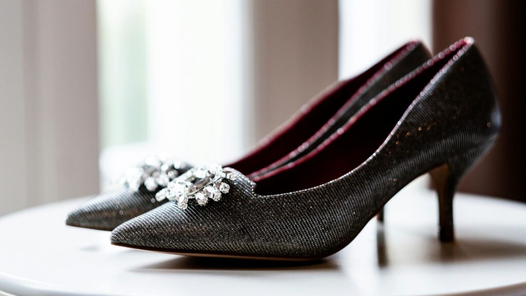 A pair of elegant, glittery black high-heeled shoes with pointed toes and crystal embellishments on the front, placed on a white round table near a softly lit window.