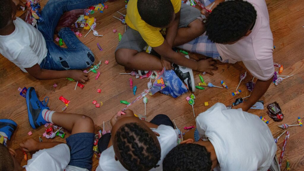 A group of children sit on a wooden floor, surrounded by scattered pieces of colorful candy. They appear to be sorting or collecting the candy, with some holding pieces in their hands. The children are dressed casually, and the scene suggests a festive or party atmosphere.
