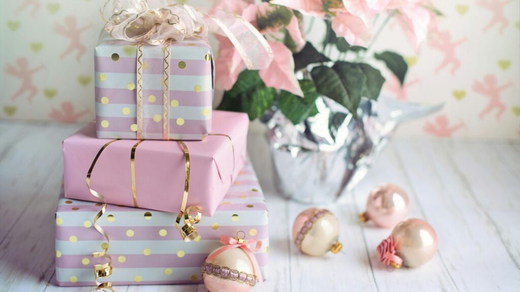 A stack of pastel-colored gift boxes wrapped in pink and lavender paper with gold polka dots and ribbons sits on a white wooden surface. Pink and white Christmas ornaments are scattered around the gifts, and a potted plant with soft pink flowers wrapped in metallic silver foil is in the background. The overall setting has a soft, festive, and elegant feel.