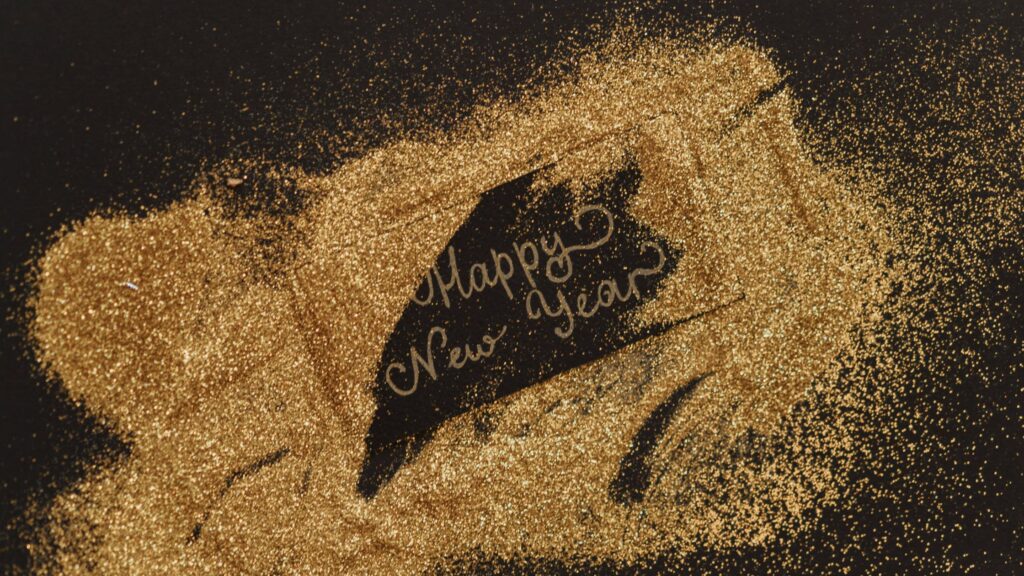 A black surface covered in scattered gold glitter reveals the handwritten message "Happy New Year" partially uncovered in the center. The shimmering glitter adds a festive and celebratory feel to the image.