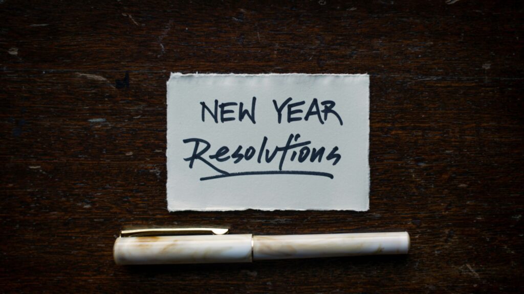 A handwritten note on torn white paper that reads 'New Year Resolutions' in bold black ink, placed on a dark wooden surface with a white and gold fountain pen lying below it.