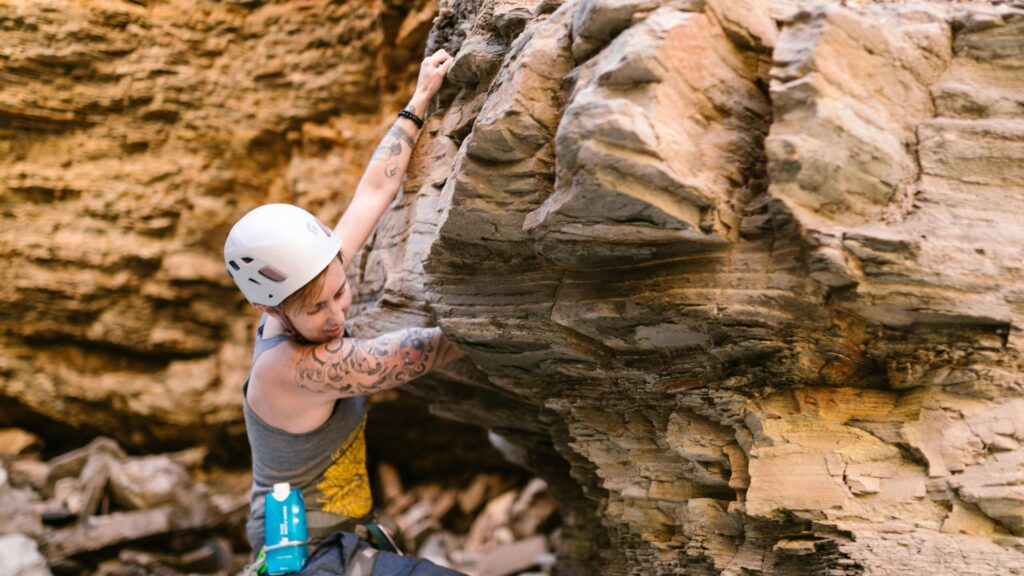 A person wearing a white helmet and gray tank top with tattoos on their arm is rock climbing on a rugged cliff face. They are gripping a protruding section of rock while looking focused, with climbing gear attached to their harness.
