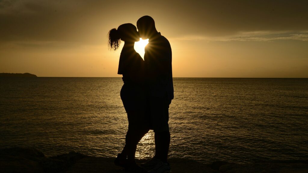 Silhouette of a couple kissing at sunset by the ocean, with the sun shining between them and calm waves in the background.