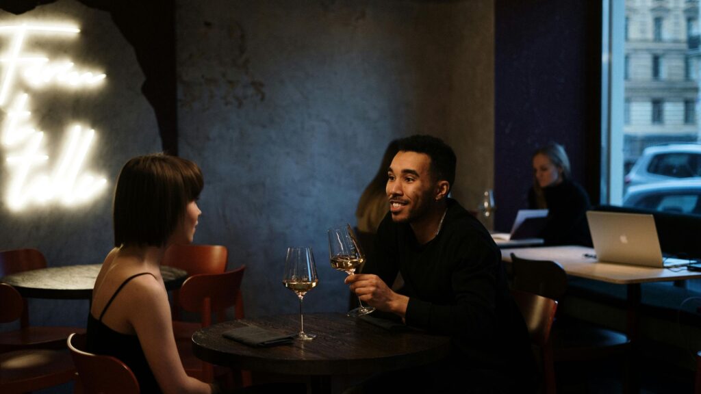 Man and woman enjoying a conversation over glasses of wine at a cozy, dimly lit café, with a neon sign and a person working on a laptop in the background.