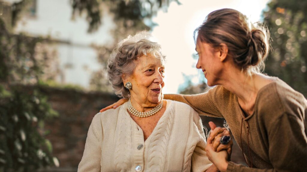 A smiling elderly woman with gray hair and pearl jewelry is warmly embraced by a younger woman with a bun, both enjoying a heartfelt moment outdoors in a sunlit garden.