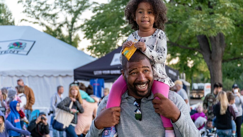 A smiling man carries a young girl on his shoulders at an outdoor event. The girl holds a snack and wears a patterned shirt and pink leggings. Tents and a crowd of people are visible in the background, suggesting a lively community gathering or festival.