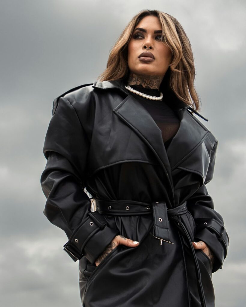 A confident woman poses against a cloudy sky, wearing a belted black leather trench coat, a pearl necklace, and a sheer top. She has long, wavy hair, bold makeup, visible tattoos on her neck and hands, and a nose ring, exuding a strong, edgy, and stylish presence.