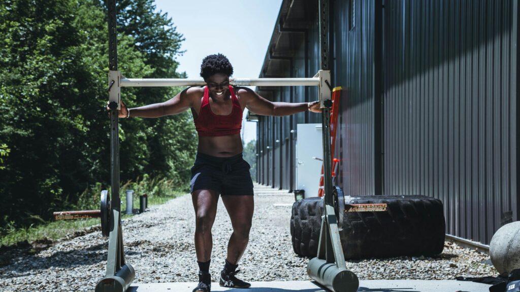 A woman wearing a red sports bra and black shorts is outdoors, lifting a heavy metal yoke with weights attached, as part of strength training. She is smiling and appears focused, with a large tire and gym equipment nearby. The setting is a gravel path between a building with metal siding and a tree-lined area, under bright daylight.