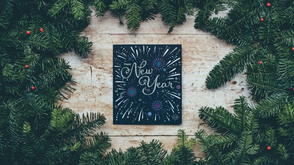 Happy New Year greeting card surrounded by green pine branches arranged in a circular wreath on a wooden background.