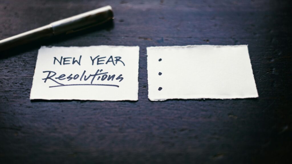 New Year resolutions written on a small paper note next to a blank checklist, with a pen on a dark wooden surface.”