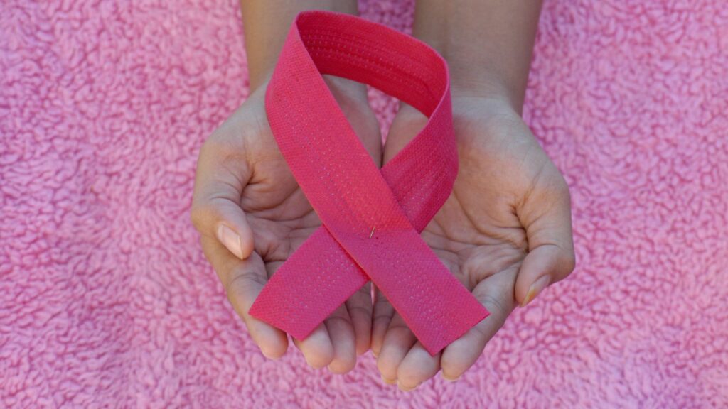 Two open hands gently holding a pink awareness ribbon against a pink textured background, symbolizing breast cancer awareness and support.