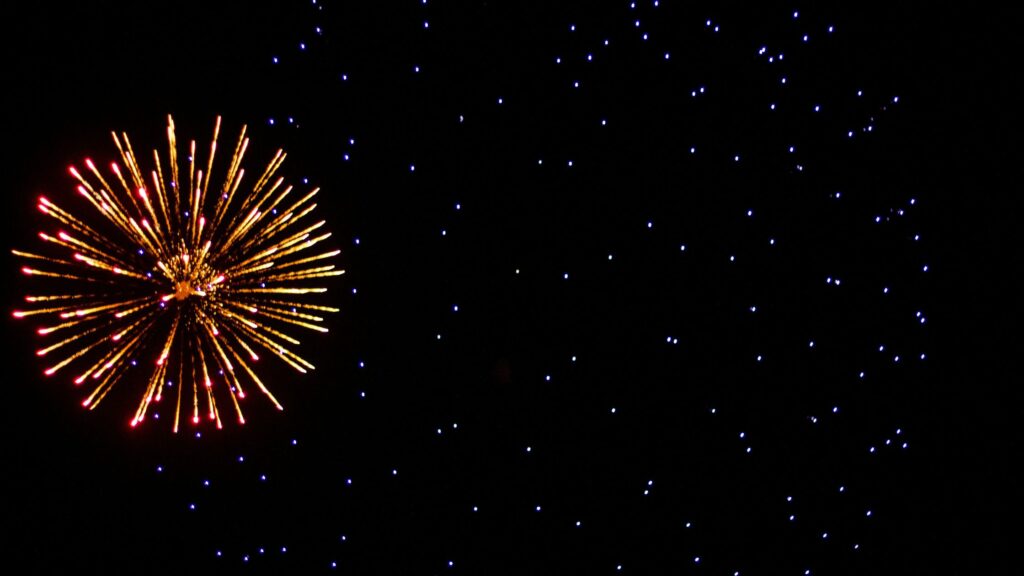A bright golden firework explodes on the left side of a dark night sky, its sparks radiating outward like a starburst, while small blue points of light are scattered across the background, resembling distant stars.