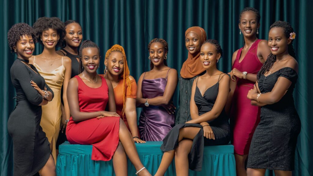 Ten young women dressed in elegant, colorful dresses pose together in front of a deep green curtain backdrop. Some are standing while others sit on a teal bench, smiling confidently at the camera. Their outfits range from red and black to gold and purple, and one woman wears a brown hijab. The group appears poised and celebratory, as if attending a formal event