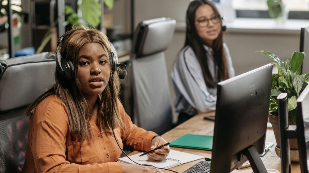 Two women working in a modern office. In the foreground, a woman wearing a headset sits at a desk with a computer, holding a pen over a notebook and looking toward the camera. In the background, another woman wearing a headset works at a computer. The workspace includes office chairs, desks, and green plants, suggesting a customer support or call center environment.