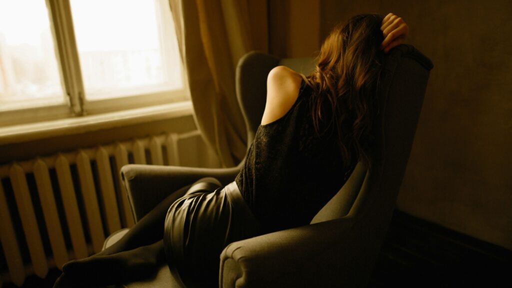 A woman slumps sideways in a cushioned armchair near a window, her face turned away, one arm resting on her head, dressed in black as dim, warm light fills the room, creating a somber, withdrawn atmosphere.