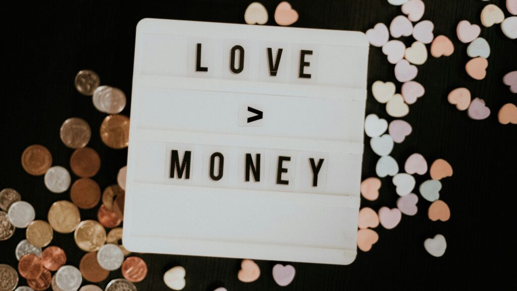 A white lightbox sign reading “LOVE > MONEY” sits on a dark surface, with scattered coins on the left and pastel heart-shaped confetti on the right.