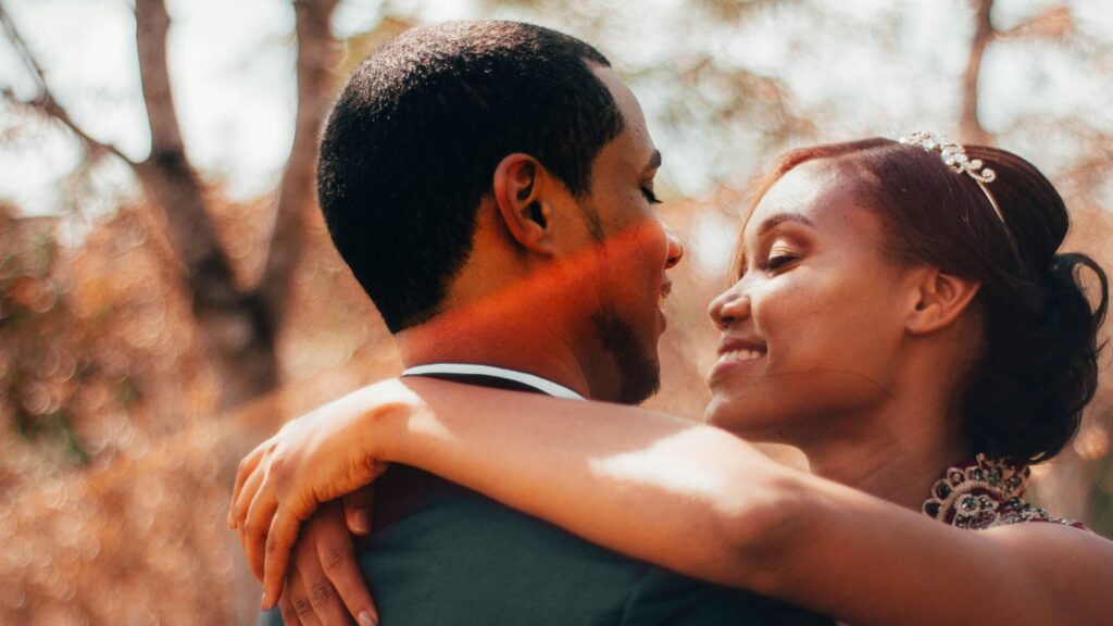 A bride and groom embrace closely outdoors, smiling at each other during a wedding photoshoot, with soft sunlight and blurred trees in the background.