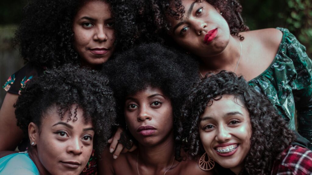 Five young women with natural curly and afro-textured hair pose closely together outdoors, their heads leaning toward one another. They display a range of expressions, from soft smiles to calm, serious looks. One woman in front smiles broadly, showing braces, while another wears bold red lipstick. The group appears relaxed and connected, framed by greenery in the background.