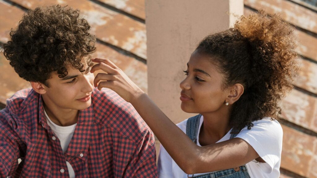 A teenage girl with curly hair tied in a puff gently adjusts a teenage boy’s curls as they sit close together on outdoor wooden steps, both smiling softly at each other in warm sunlight.