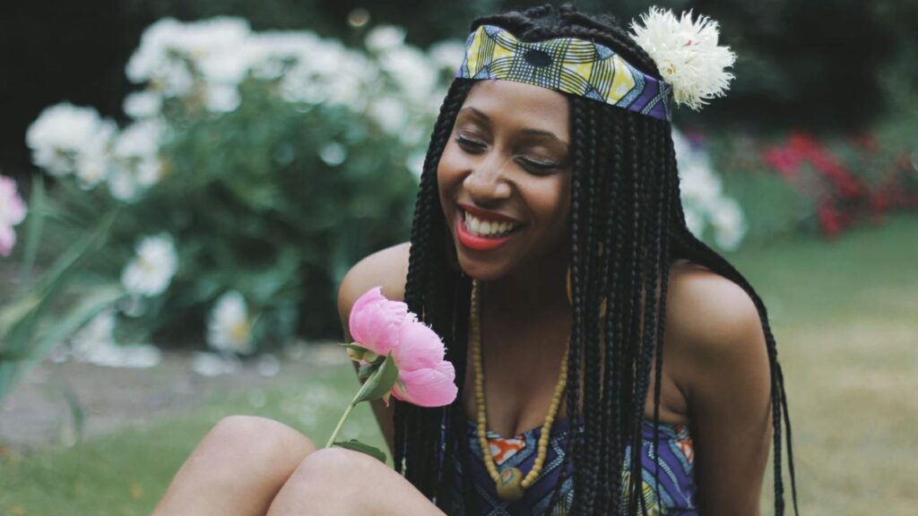 A smiling woman with long braided hair wears a colorful patterned headband and matching outfit while holding a pink flower in a lush garden, with soft greenery and white blooms blurred in the background.