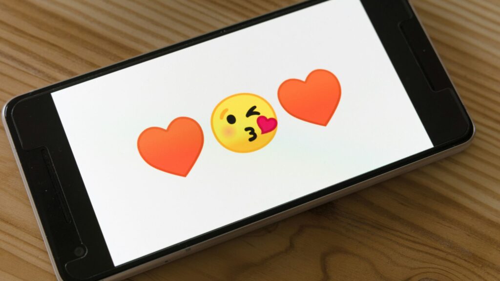 Smartphone lying horizontally on a wooden table, displaying a white screen with a winking face blowing a kiss emoji in the center, flanked by two red heart icons on either side.