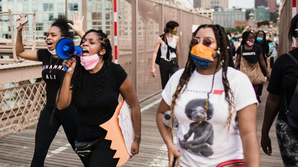 Group of women marching across a city bridge during a protest, with one woman shouting into a megaphone while others raise their hands and walk alongside her. Several participants wear face masks, and buildings are visible in the background.
