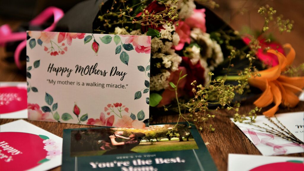 A floral Mother's Day greeting card sits on a wooden table beside a bouquet of flowers. The card, decorated with pink flowers and green leaves, reads “Happy Mothers Day” and includes the quote, “My mother is a walking miracle.” Surrounding the card are additional Mother's Day cards, ribbons, and a partially visible photograph, creating a warm and celebratory scene filled with soft colors and floral arrangements.