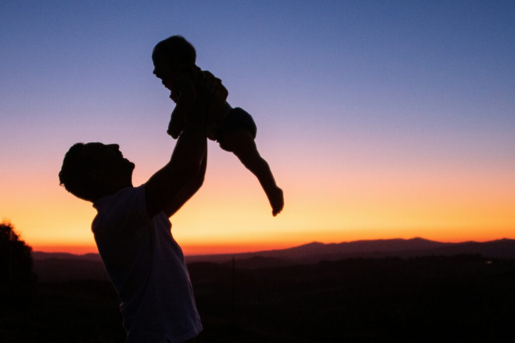 Silhouette of an adult lifting a baby into the air against a colorful sunset sky. The horizon glows with warm shades of orange, pink, and purple, while distant mountains form a dark outline in the background. The scene conveys joy, affection, and a tender parent-child moment.