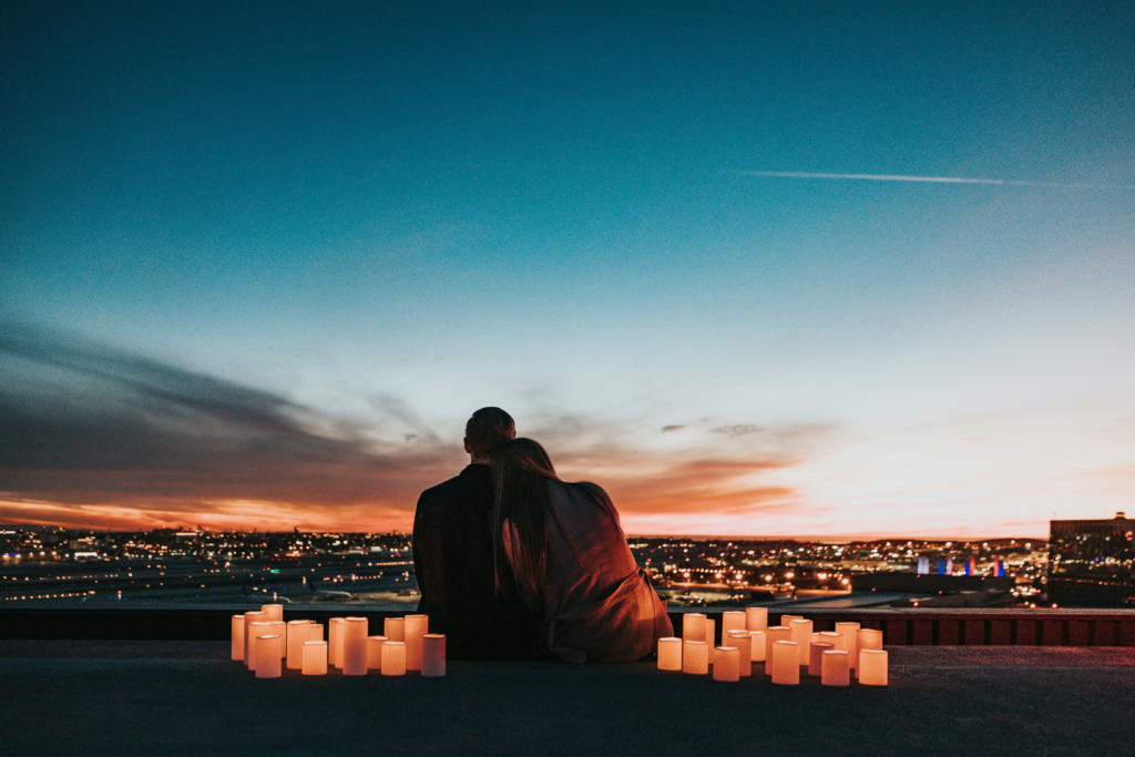 A couple sits closely together on a rooftop at dusk, overlooking a city illuminated by thousands of lights. The person on the right rests their head on the other's shoulder as they watch the sunset fade into a deep blue sky. Dozens of glowing candles line the edge of the rooftop in front of them, creating a warm, intimate atmosphere against the expansive urban skyline.