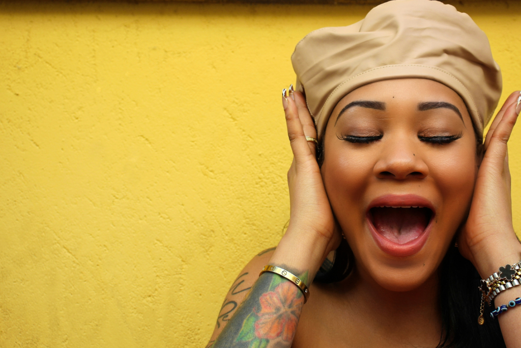 A close-up portrait of a woman against a textured yellow wall, wearing a beige satin head wrap with her eyes closed and mouth open in an expressive, joyful pose. She gently holds the sides of her face with both hands, showing gold rings, bracelets, and manicured nails. A colorful floral tattoo is visible on her forearm, creating a vibrant, energetic composition with ample yellow negative space on the left.