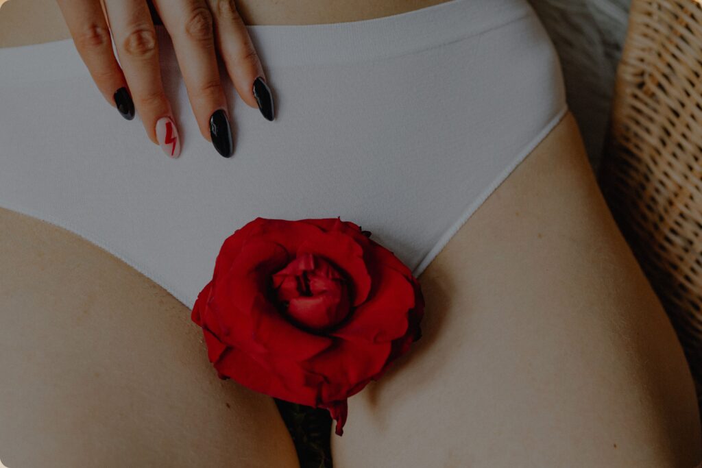 A close-up of a person wearing white underwear with a red rose placed over the pelvic area. One hand rests gently on the waistband, featuring glossy black nails and a white accent nail with a red lightning bolt design. The image uses soft, warm lighting and is styled as an artistic, body-positive composition symbolizing femininity, menstruation, or reproductive health.