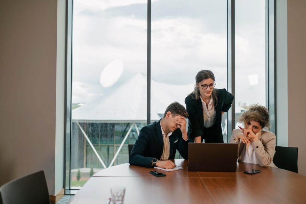 Three professionals gather around a laptop in a modern conference room during what appears to be a challenging meeting. One person sits with their head in their hand, another leans over the table reviewing the screen, and a third rests their chin on their hand while holding a pen, suggesting deep thought or concern. Large floor-to-ceiling windows behind them let in natural light, with a cloudy sky and buildings visible outside. Smartphones, a notebook, and a laptop sit on the wooden meeting table, reinforcing the workplace setting.