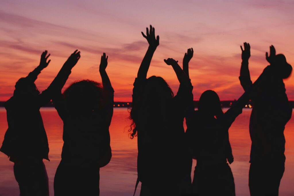 Five silhouetted figures stand together along a waterfront at sunset, raising their hands toward the colorful evening sky. The glowing orange, pink, and purple hues reflect on the water, conveying a sense of connection, celebration, and togetherness.