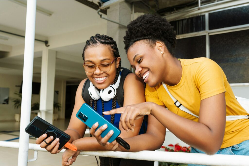Two young Black women smiling together while looking at a smartphone. One woman holds the phone while the other points at the screen, as they lean against a railing outdoors.
