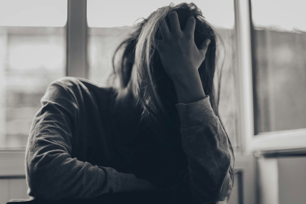 A woman sits by a window with her head resting in her hand, appearing overwhelmed or distressed, in a muted black-and-white setting.