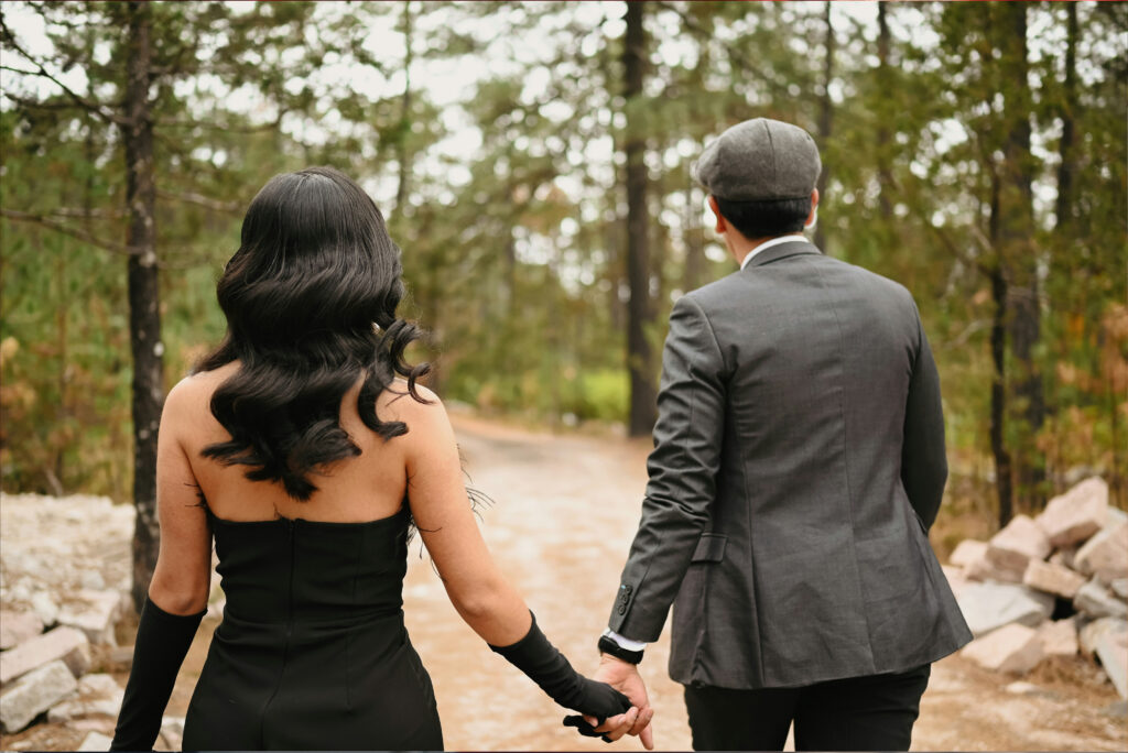 A couple holding hands as they walk along a wooded path, viewed from behind. The woman wears a black dress with long gloves, while the man wears a gray suit and flat cap.