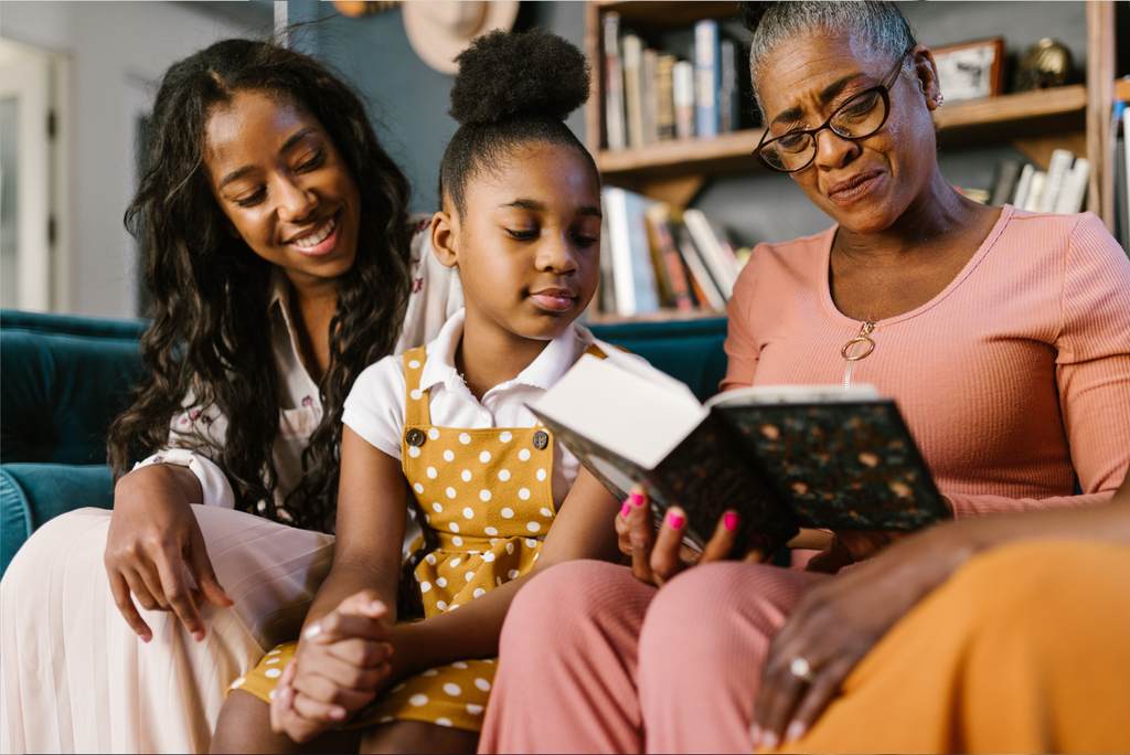 Three generations of Black women sitting together on a couch, smiling and reading a book in a warm, cozy home setting.
