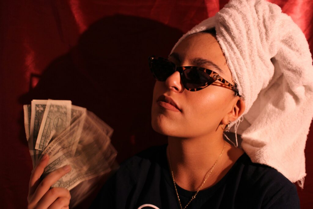 A woman wearing sunglasses and a white towel wrapped around her head fans herself with a stack of U.S. dollar bills against a dark red background.